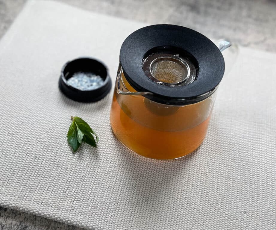 A pot of green tea hair rinse sits next to a green tea leaf.