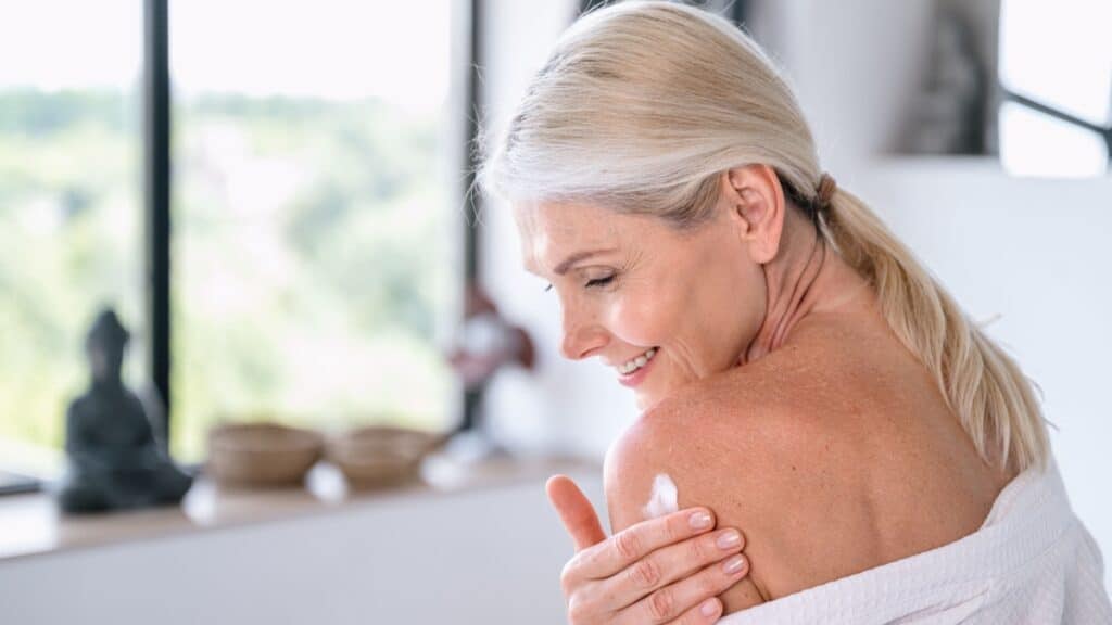 Mature woman in white bathrobe applying moisturizer cream lotion on shoulder