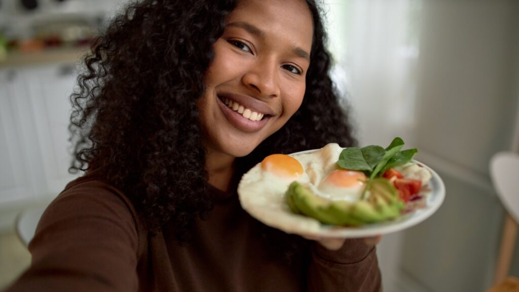 smiling woman showing her colorful delicious and healthy breakfast with eggs, avocado and greens