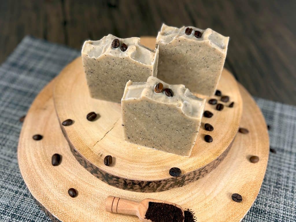 Three bars of coffee soap sit ontop a set of wooden boards, with coffee beans surrounding it