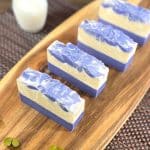 Four bars of lavender goats milk soap sit on a wooden board with a glass of milk sat next to it