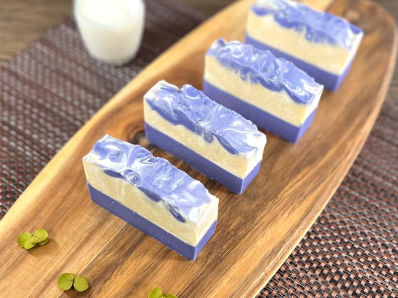 Four bars of lavender goats milk soap sit on a wooden board with a glass of milk sat next to it