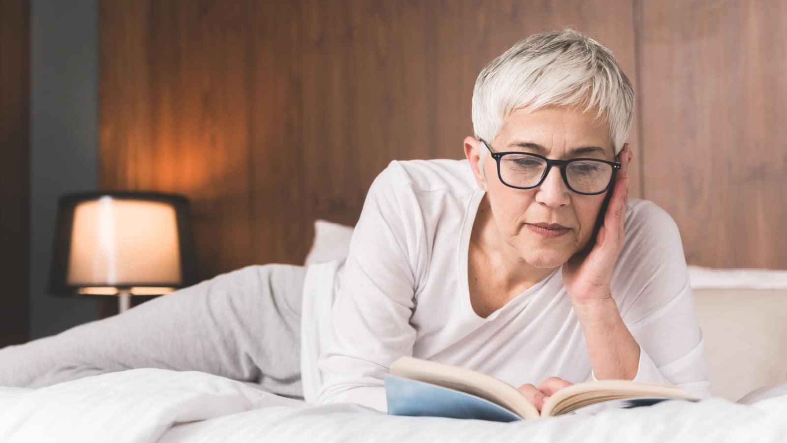 mature woman with glasses lying on a comfy bed and reading a book mature woman with glasses lying on a comfy bed and reading a book