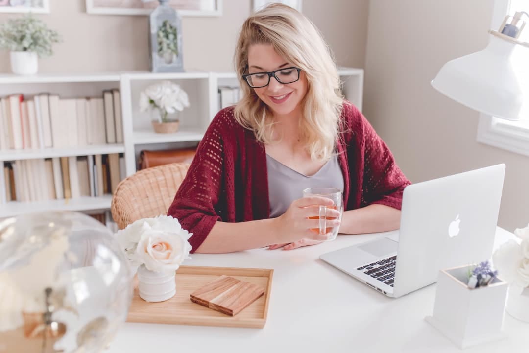 Photo by Paige Cody woman smiling holding glass mug sitting beside table with MacBook